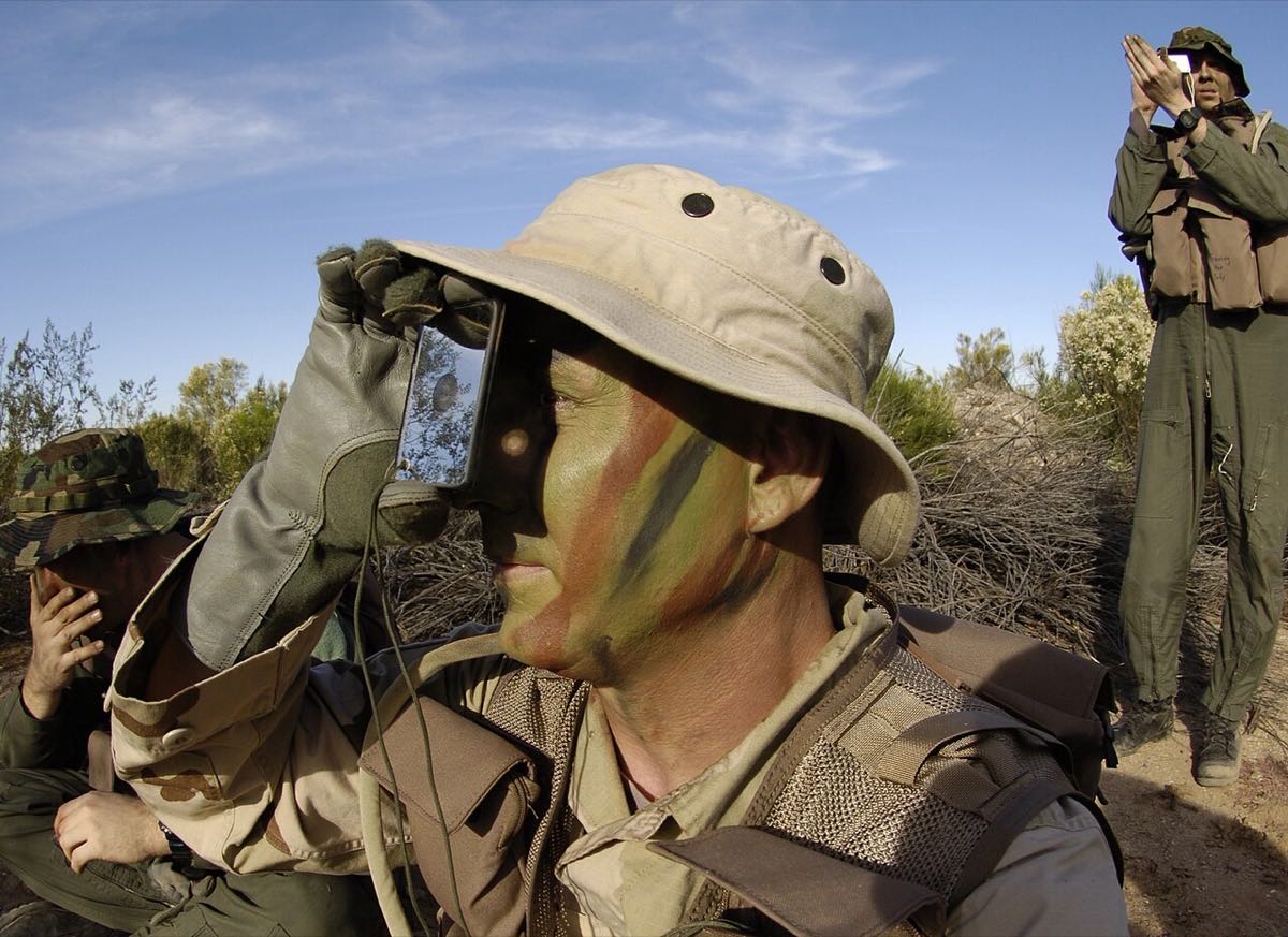 Survival trainee holding a glass signal mirror at eye level and sighting toward a target, another trainee doing the same in the background